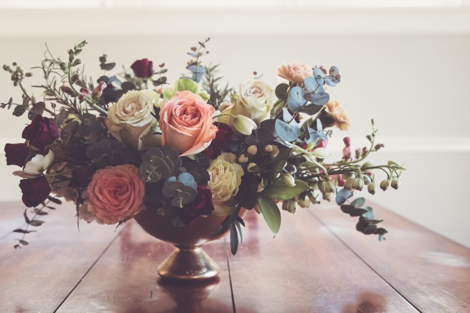 A brass footed bowl holding roses, eucalyptus and seasonal blooms on a wooden table