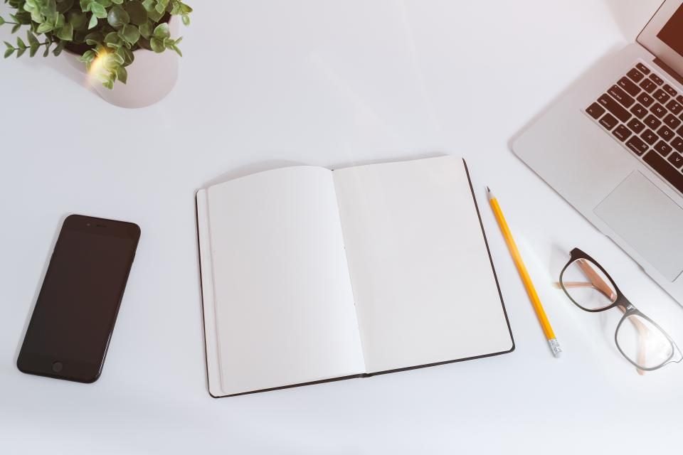 An open blank notebook, pencil and reading glasses on a pale desk beside a laptop and a small plant