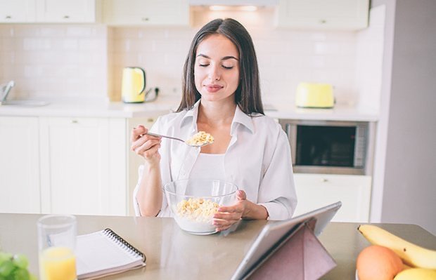 A woman eating a bowl of cereal at a kitchen counter beside a tablet and notebook